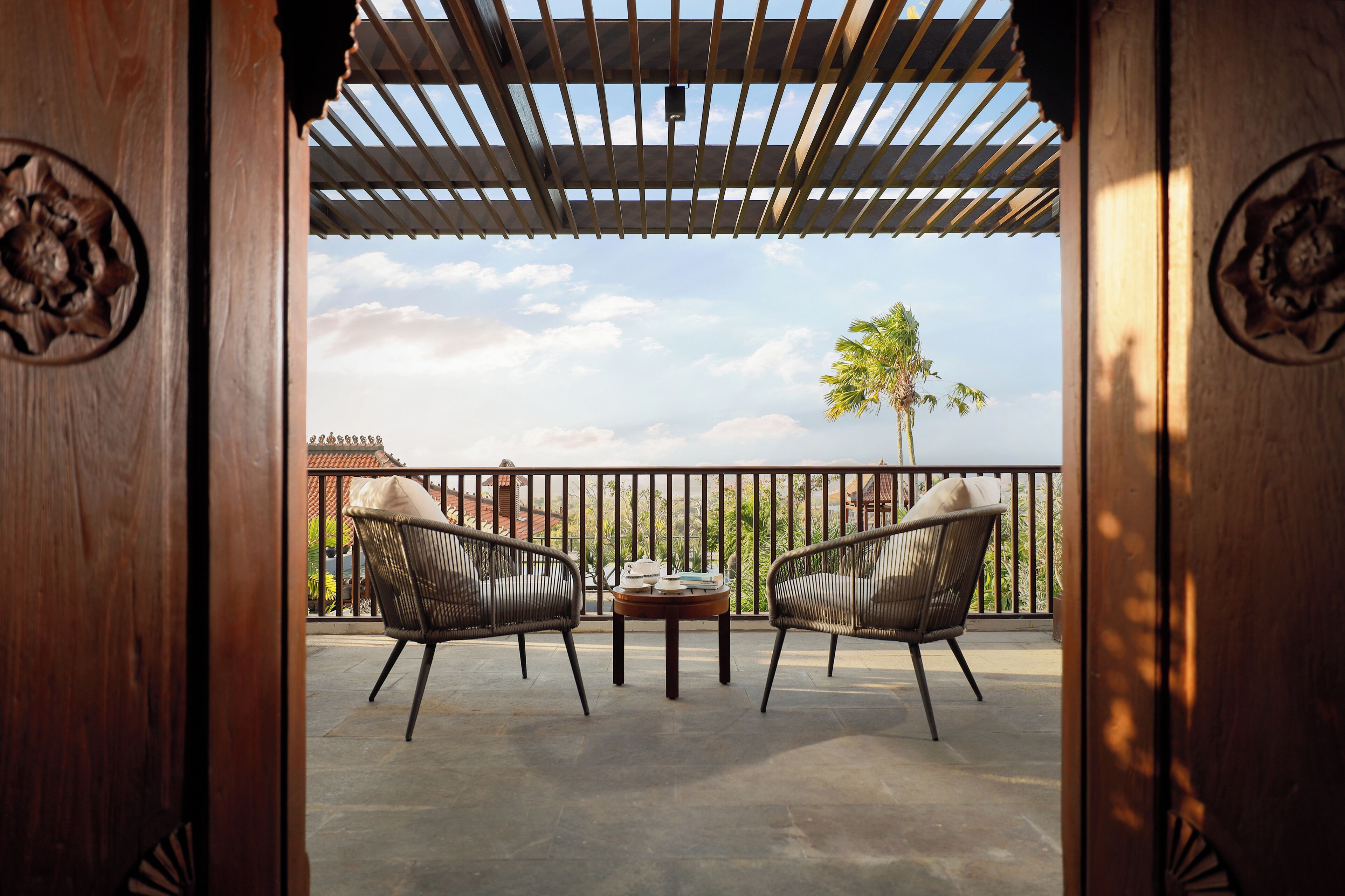 A balcony with two wicker chairs, a small table, and a view of a palm tree and buildings.