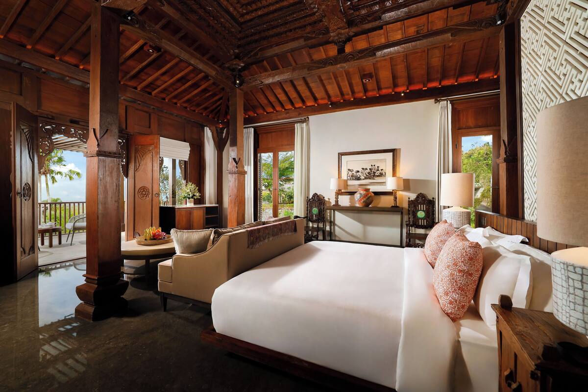Une chambre spacieuse avec un grand lit, un coin salon avec un canapé et des fauteuils, un plafond en bois et une vue sur l’extérieur.