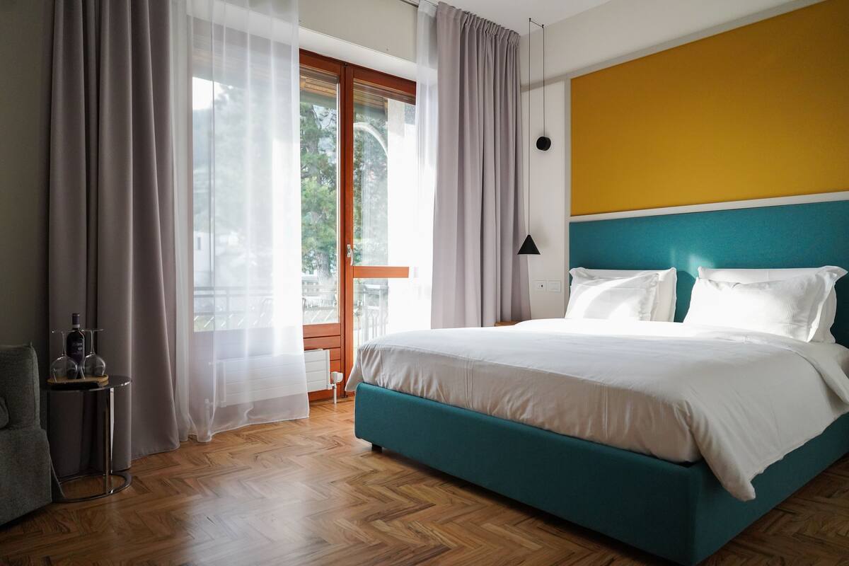 Un dormitorio moderno con una cama verde azulada, ropa de cama blanca y vistas al exterior a través de cortinas translúcidas.