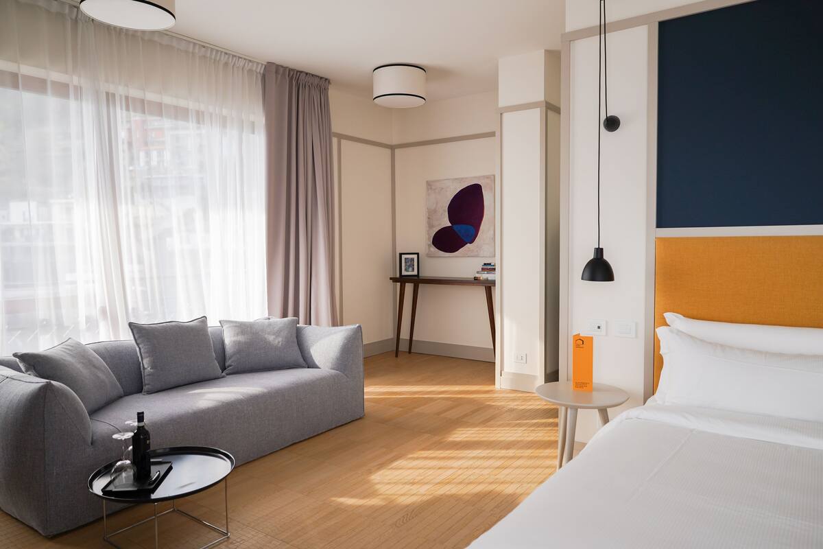 Una habitación de hotel moderna con un sofá gris, una mesita redonda con una botella, una cama con ropa blanca, un escritorio con una lámpara y un cuadro abstracto grande en la pared.