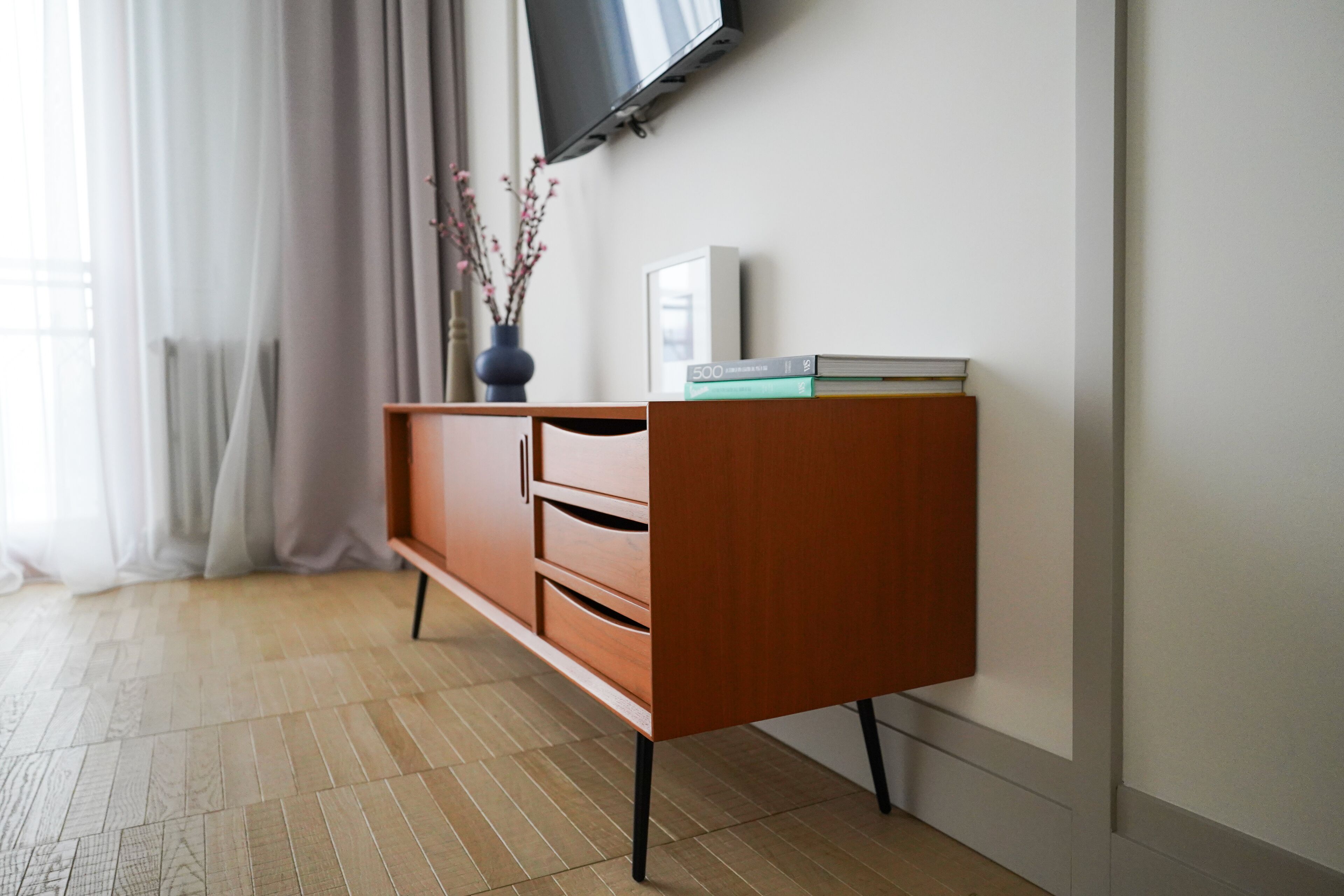 A modern living room with a wooden cabinet, a flat-screen TV mounted on the wall, and a vase with branches on a table.