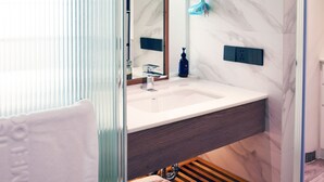 A modern bathroom with a glass shower enclosure, a marble wall, a wooden shelf with towels, and a large mirror. - Studio - Serviced Apartment Sea View - Isla Melocotón Ven67 Phu Quoc (Thành phố Phú Quốc)