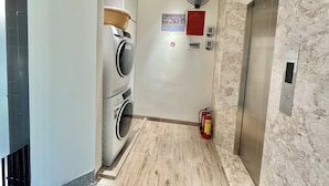 A laundry area with stacked washing machines, a fire extinguisher, and a wall-mounted fan. - Studio - Serviced Apartment Sea View - Isla Melocotón Ven67 Phu Quoc (Thành phố Phú Quốc)