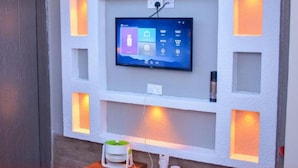 A wall-mounted TV with a control panel, recessed lighting, and a small device on a shelf below.