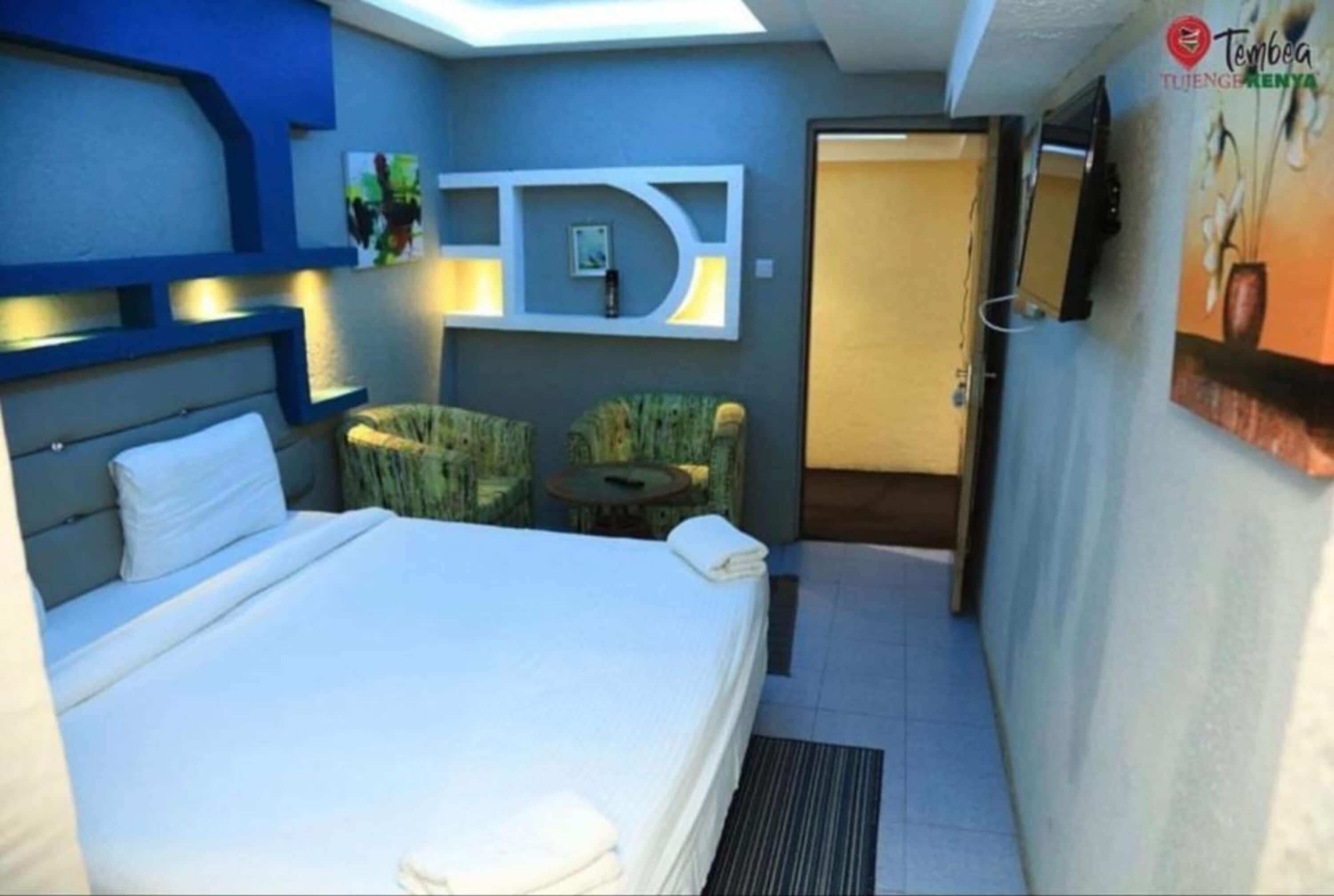 Habitación de hotel con cama, un pequeño rincón de estar con dos sillas, un televisor montado en la pared y una puerta que da a otra habitación.