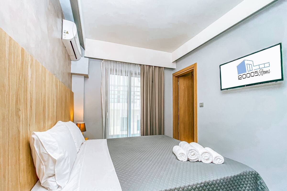 A hotel room with a bed, a bedside table, a lamp, a window with curtains, a wall-mounted air conditioner, and a sign indicating the room type.