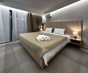 A modern hotel room with a large bed, a bedside table with a lamp, and a view of the outside through a window with curtains. - RodosRooms (Rhodes)
