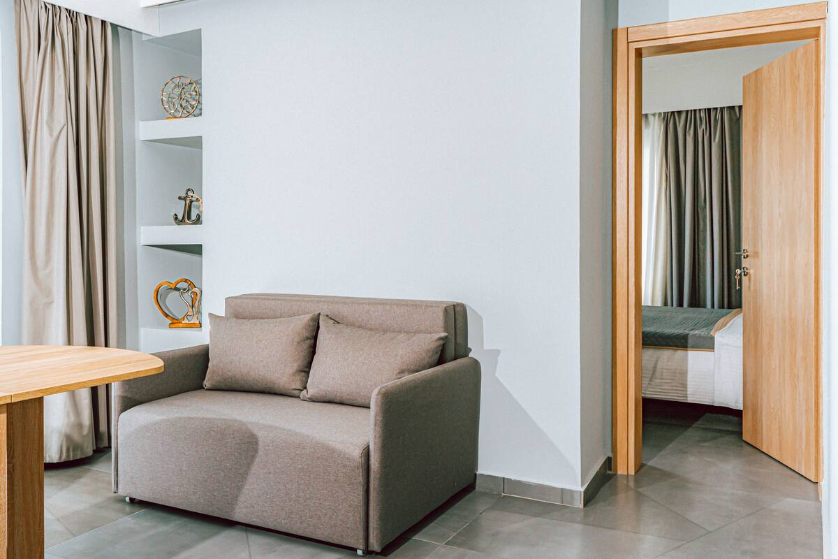 A modern room with a beige sofa, a wooden table, and a bed in the background.