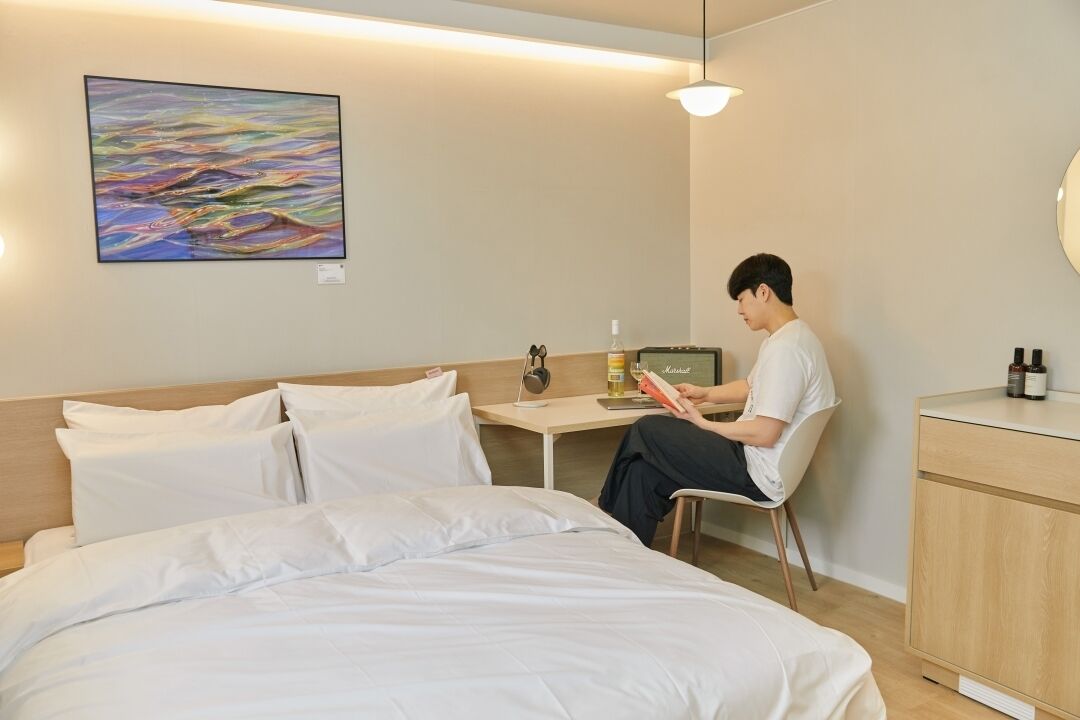 A hotel room with a bed, a desk with a laptop, a chair, a bedside table with a bottle, and a painting on the wall.