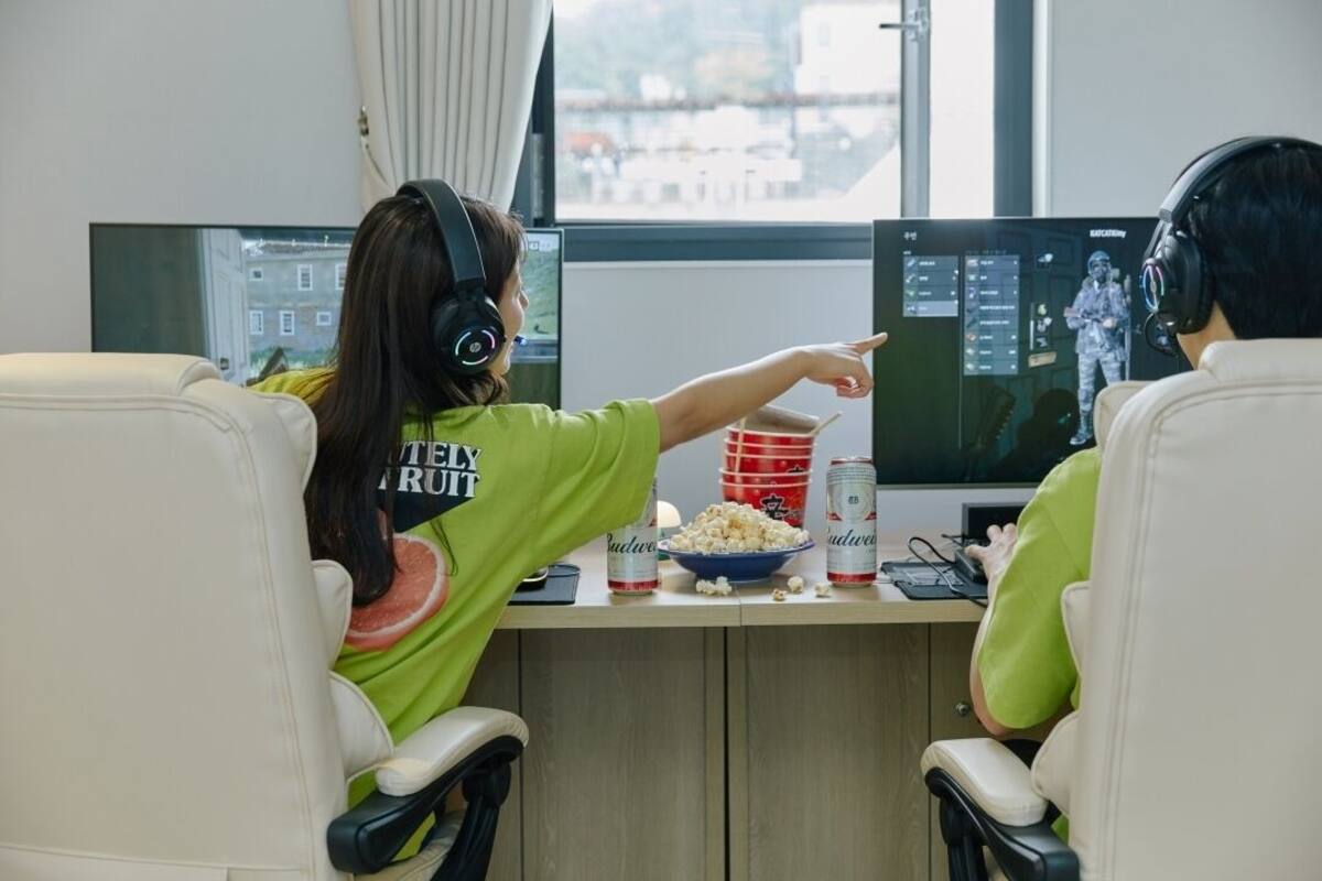 Two people sitting at a desk with computer monitors, a bowl of popcorn, and two cans of beer.