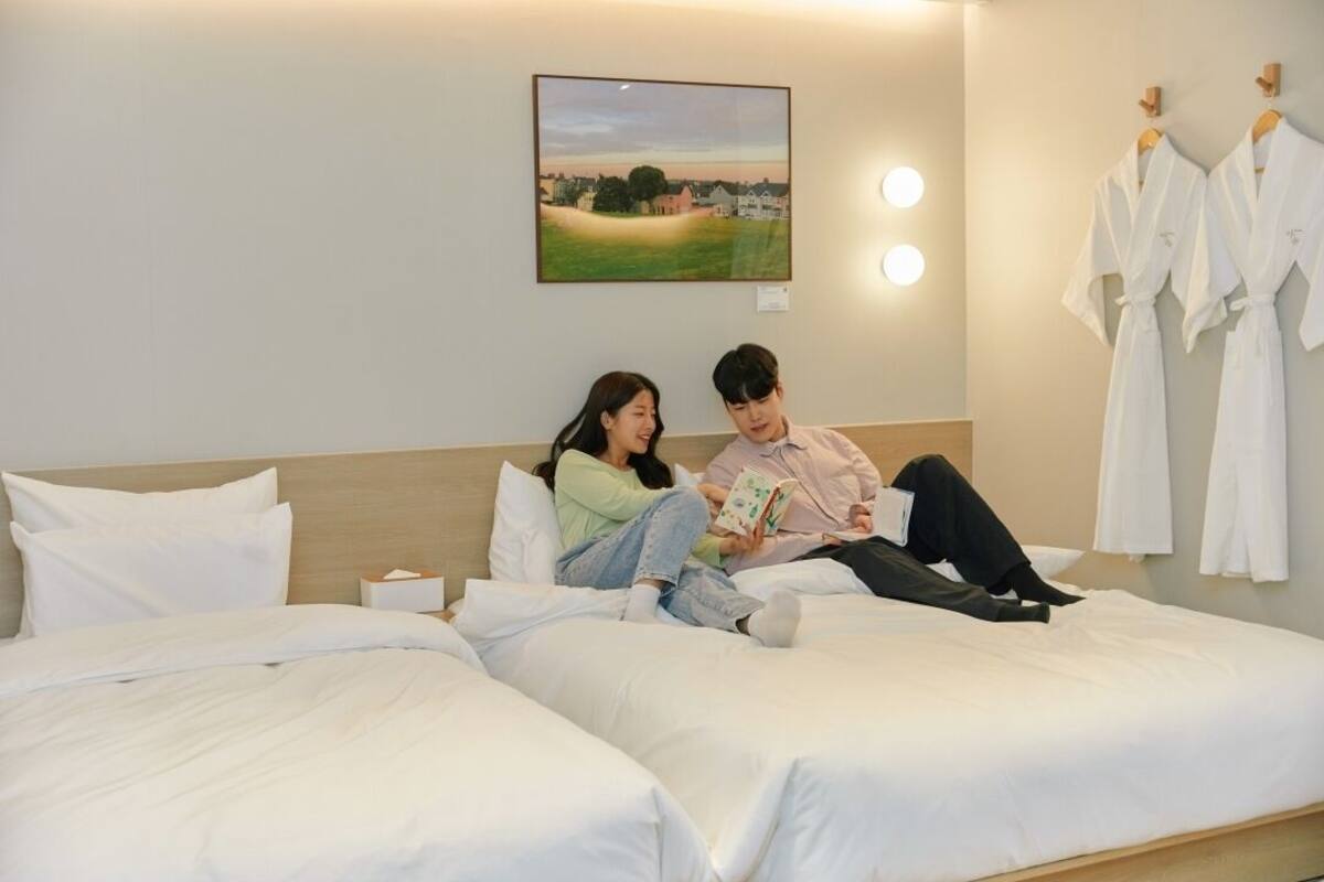 A hotel room with two beds, a painting on the wall, and white robes hanging on the wall.