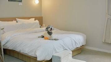 A neatly made bed with white linens, a small dog wearing a vest, and a painting on the wall.