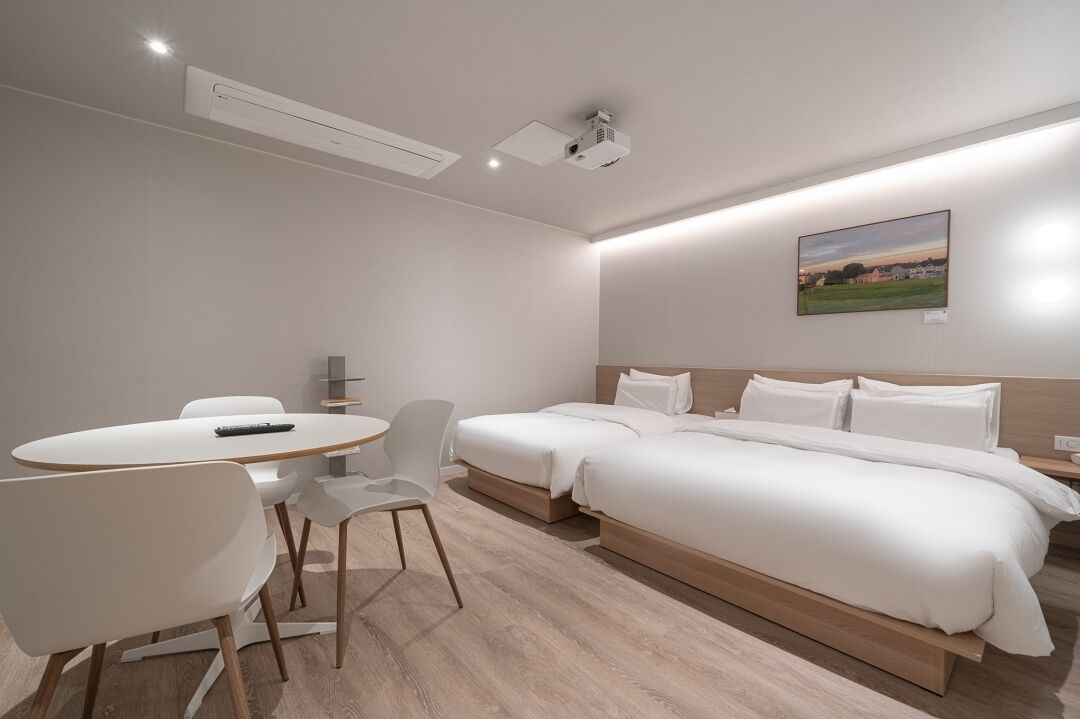 A modern hotel room with a single bed, a small table with chairs, a projector, and a painting on the wall.