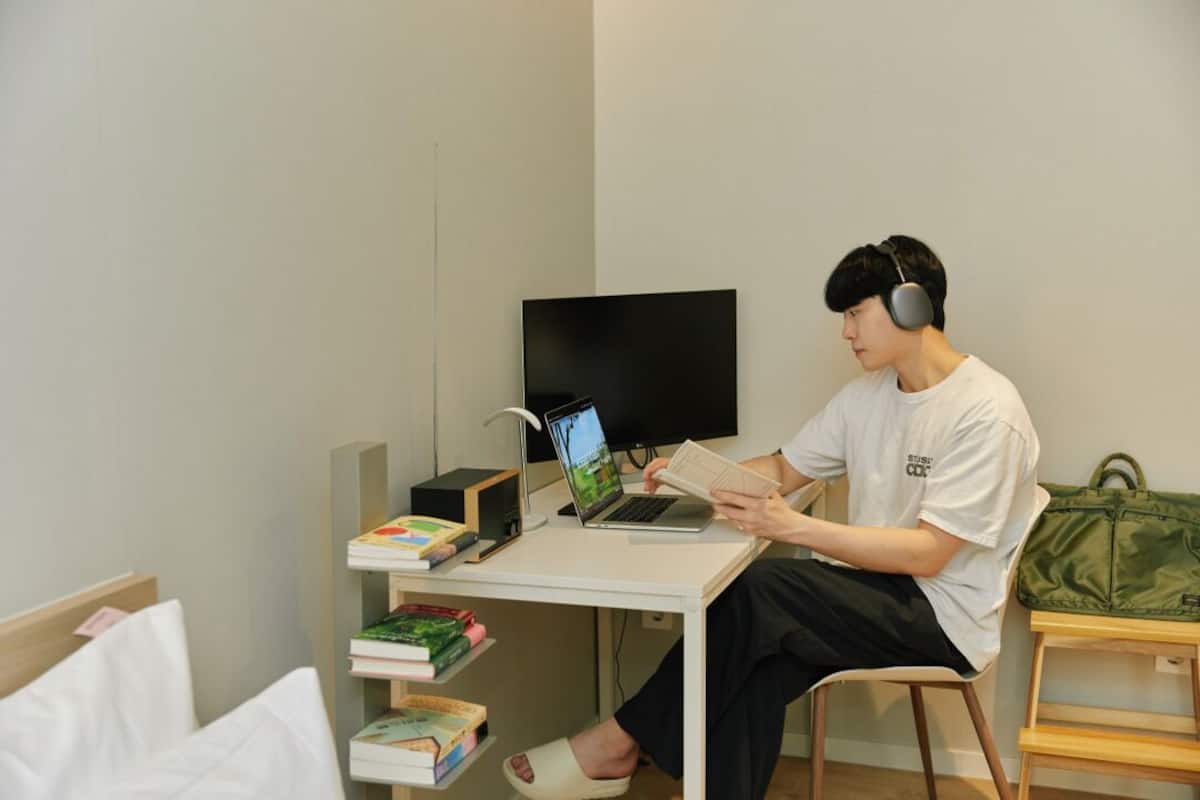 A person sitting at a desk with a laptop and books, headphones on, in a room with a flat-screen TV and a bookshelf.