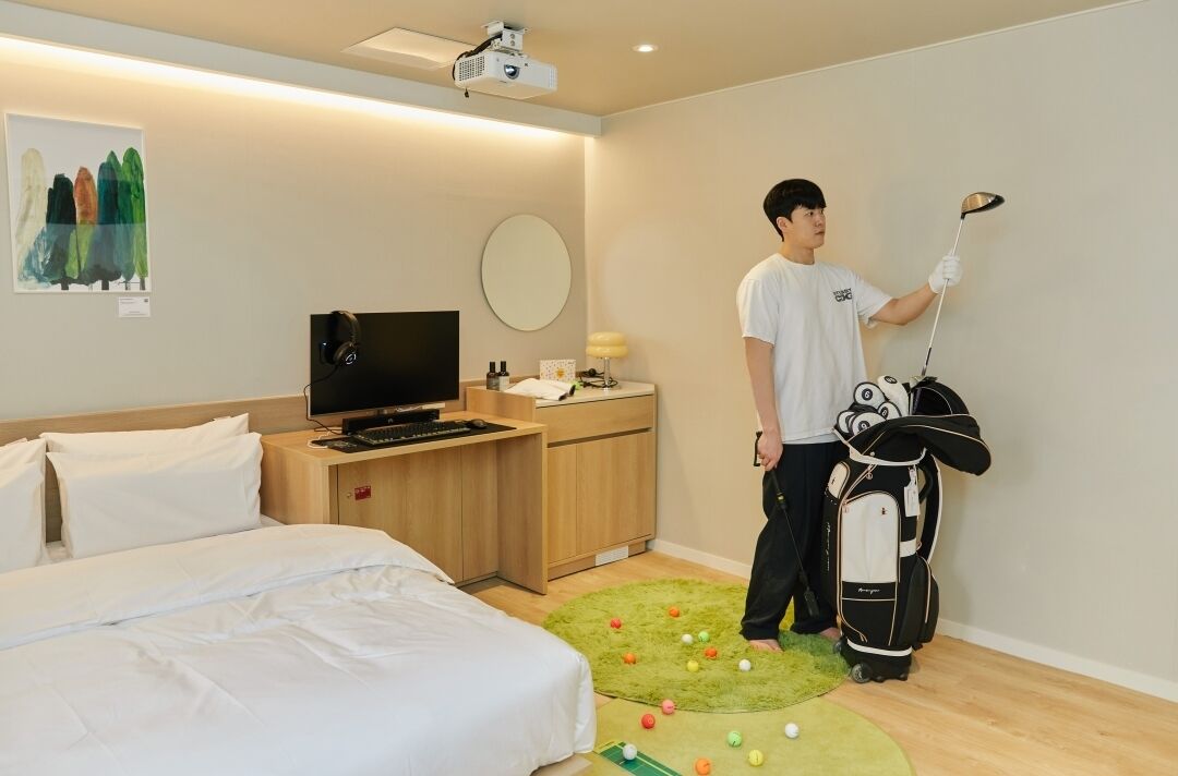Quarto de hotel com cama, escrivaninha com televisão, mala de golfe e tapete verde com bolas de golfe.