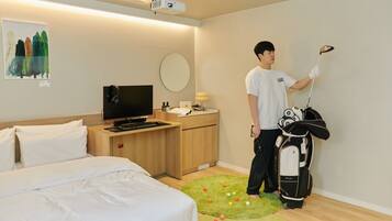 Quarto de hotel com cama, escrivaninha com televisão, mala de golfe e tapete verde com bolas de golfe.