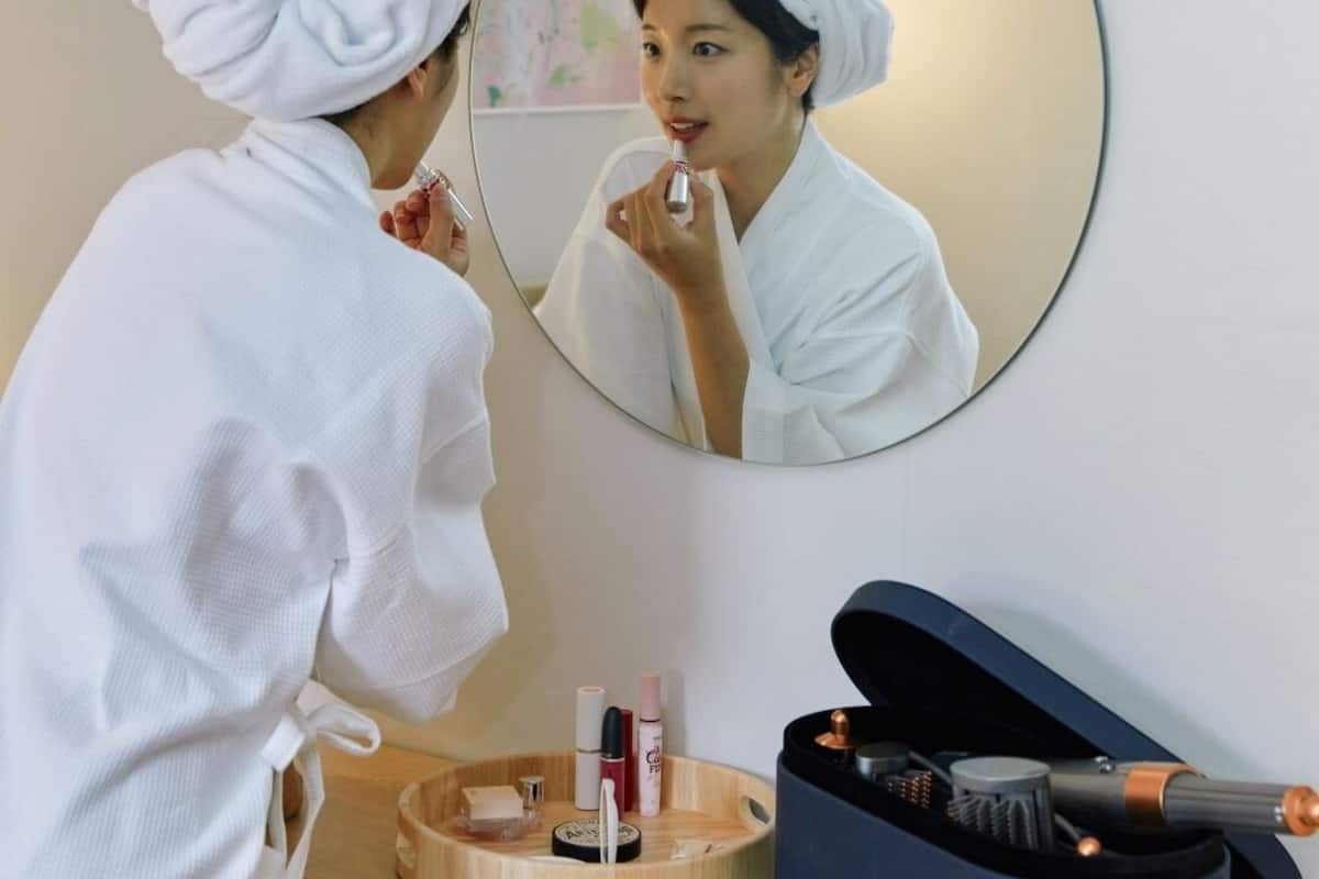 A person in a white robe and towel head wrap standing in front of a mirror, applying lipstick. A round mirror is mounted on the wall, and a wooden tray with various cosmetic items is on the counter.