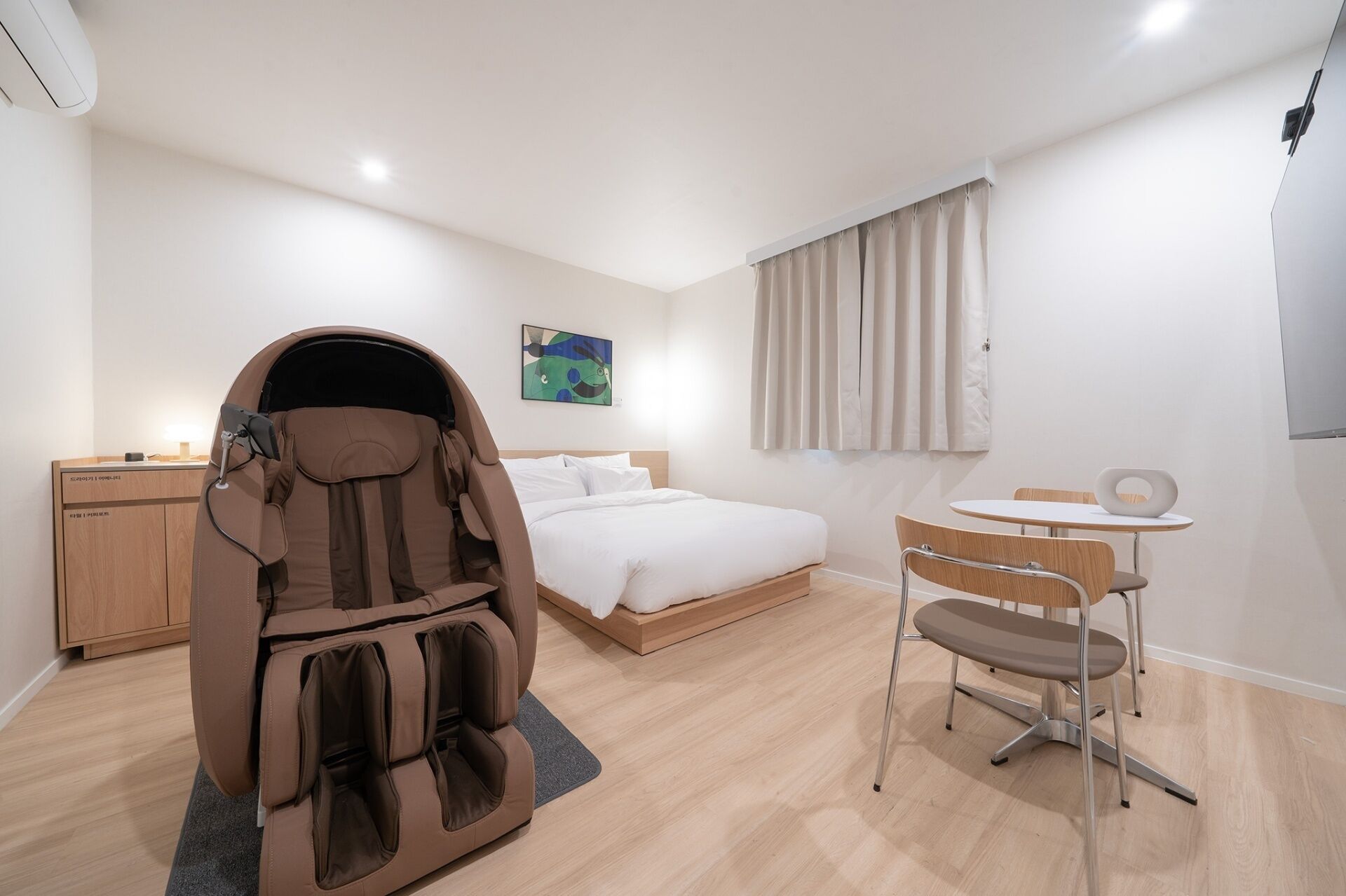 A modern hotel room with a massage chair, a bed, a desk, and a small table with chairs.