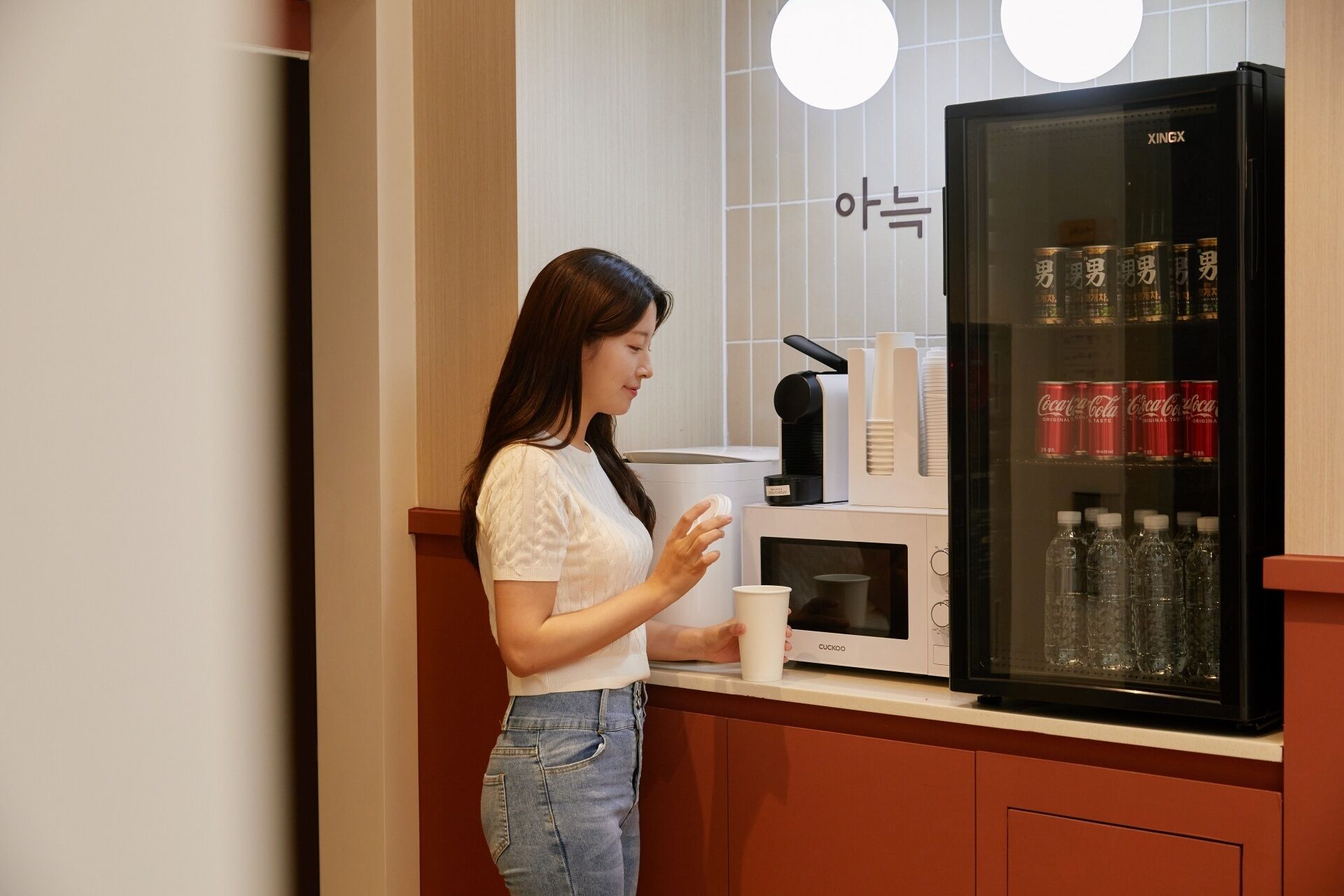 A person standing in a kitchen with a refrigerator stocked with beverages, a coffee machine, and a microwave.