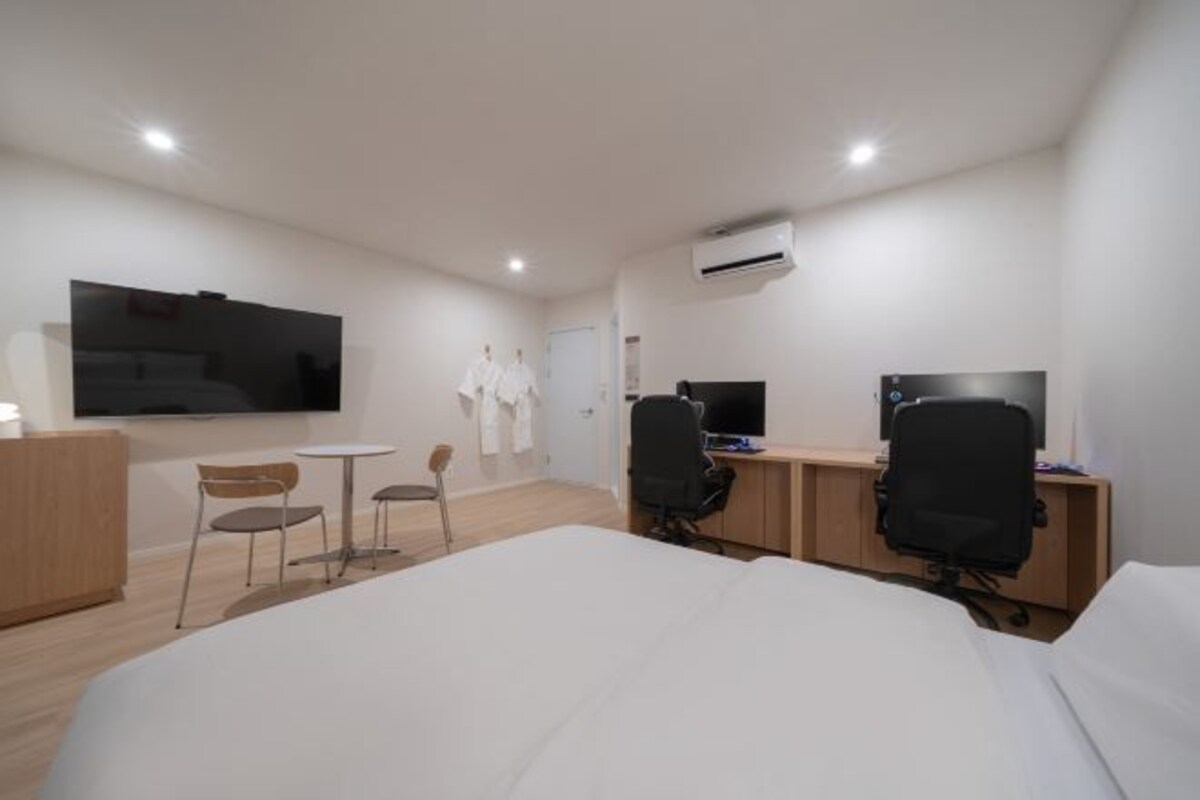 Ein Hotelzimmer mit einem Bett, zwei Schreibtischen mit Computern, einem Fernseher, einem kleinen Tisch mit Stühlen und einer Klimaanlage.