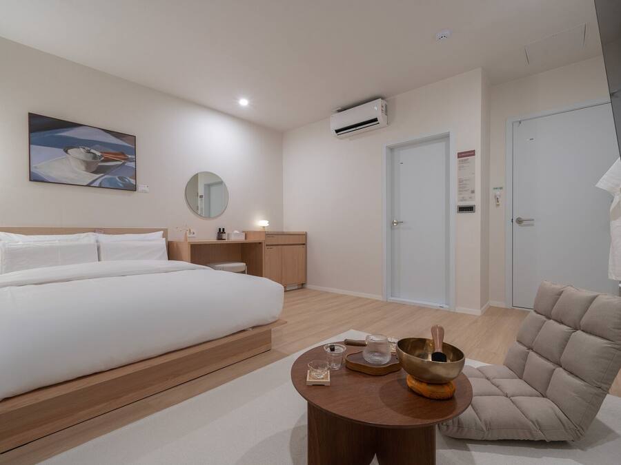 A modern hotel room with a large bed, a round mirror, a bedside table with a lamp, a small round table with a bowl and candles, and a beige sofa.