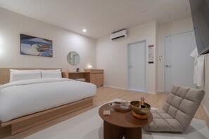Une chambre d’hôtel moderne avec un grand lit, un miroir rond, une table de chevet avec une lampe, une petite table ronde avec un bol et des bougies, et un canapé beige.