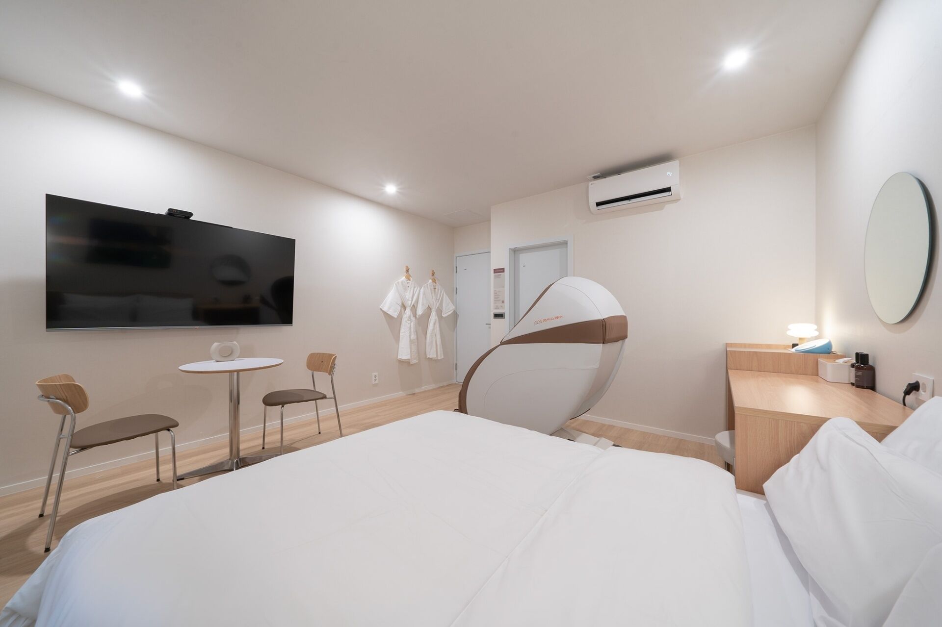 Une chambre d’hôtel moderne dotée d’un grand lit, d’un téléviseur à écran plat, d’un fauteuil massant, d’une petite table, de deux chaises, d’un bureau avec une lampe et d’un appareil de climatisation.