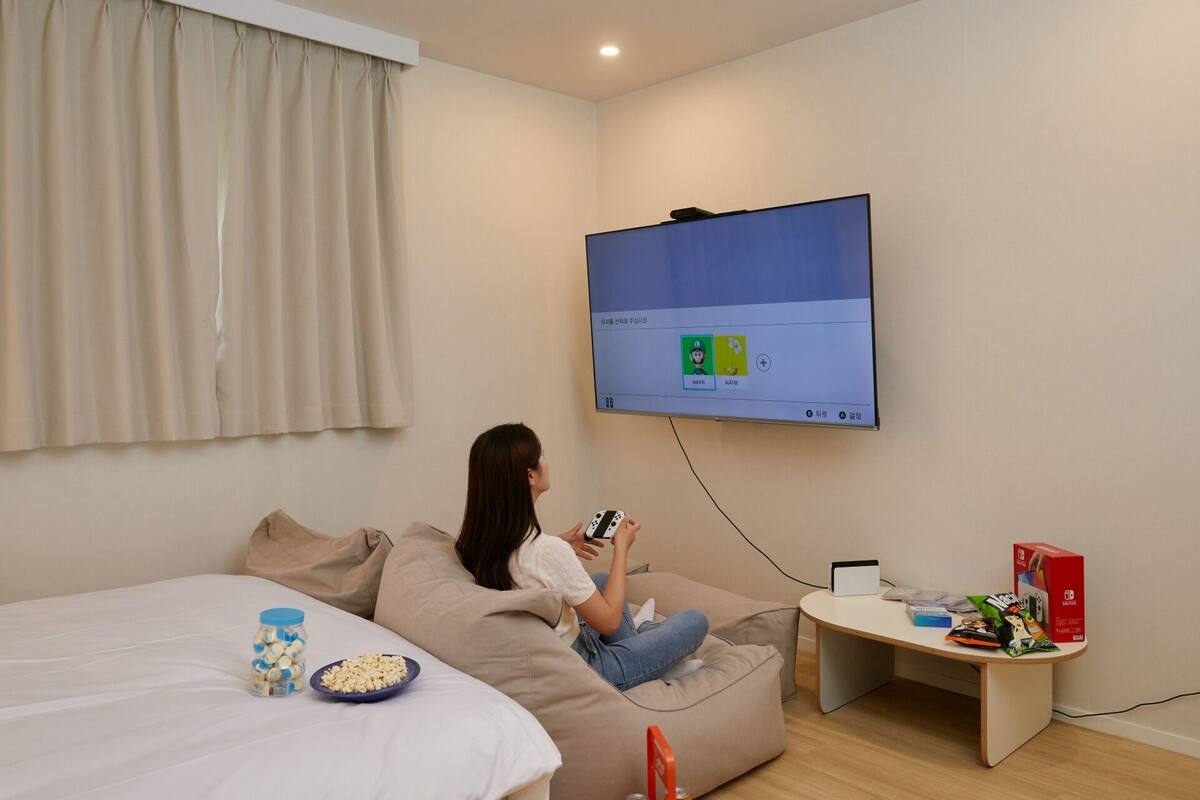 Iemand zit op een zak stoel en speelt een videospel, terwijl een aan de muur bevestigde televisie een spelinterface toont.