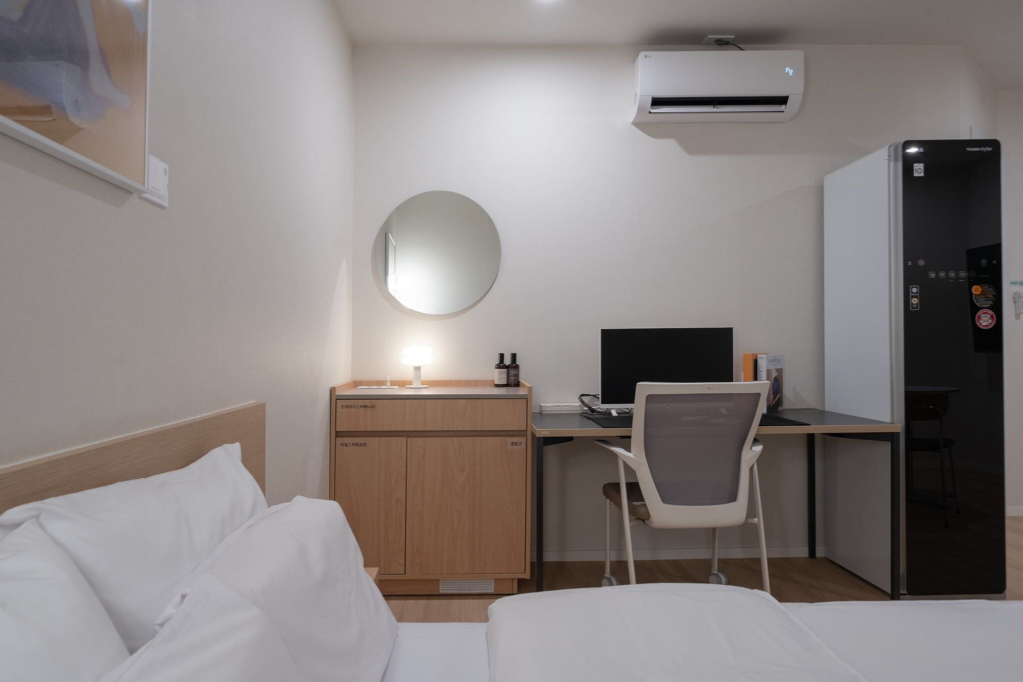 A hotel room with a bed, a desk, a chair, a mirror, a refrigerator, and an air conditioning unit.