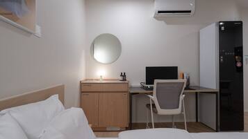 A hotel room with a bed, a desk, a chair, a mirror, a refrigerator, and an air conditioning unit.