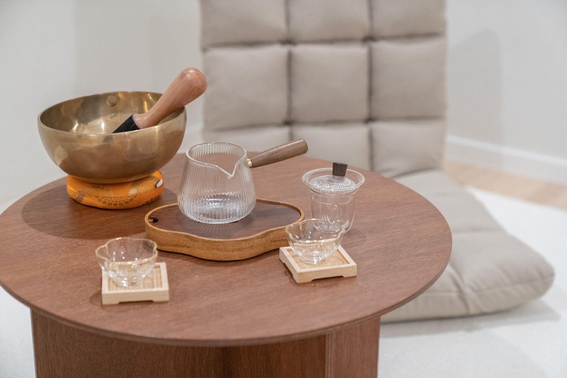 A wooden table with a brass bowl, a glass pitcher, and two glass cups on a wooden tray.