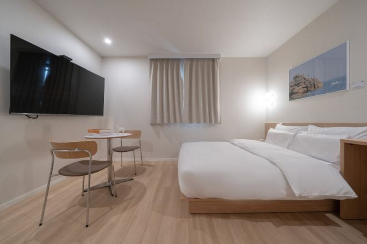 A hotel room with a bed, a TV mounted on the wall, a small table with chairs, and a window with curtains.