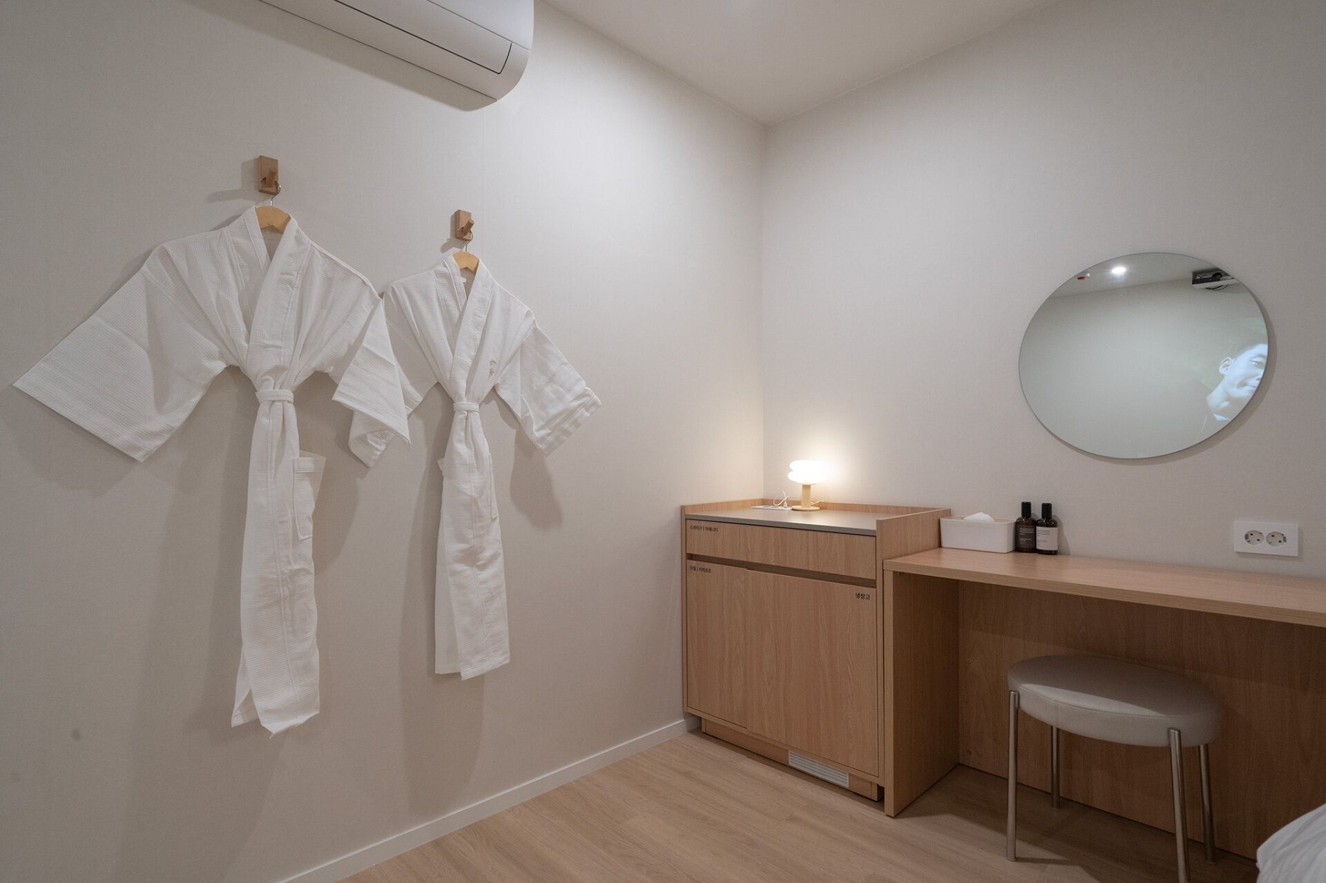 Une salle de bain avec deux peignoirs blancs suspendus à un porte-serviettes en bois, un meuble en bois avec un miroir, un petit tabouret et un climatiseur fixé au mur.