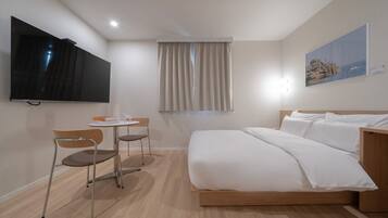 A hotel room with a bed, a desk with two chairs, a flat-screen TV, a window with curtains, and a painting on the wall.