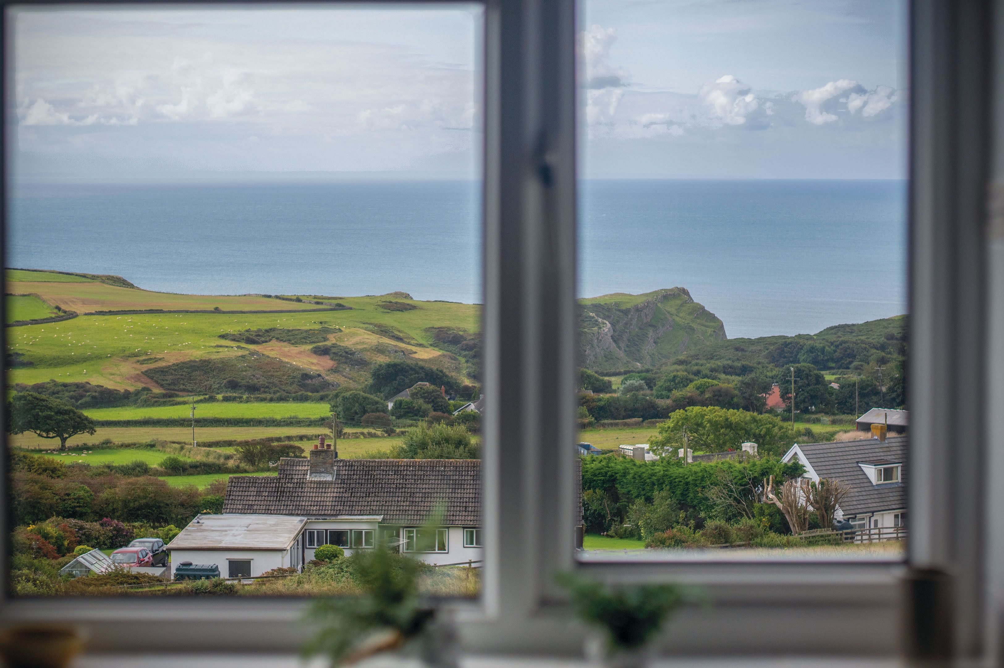 Vista costera desde una ventana, que incluye un paisaje con hierba, algunas casas y el mar.