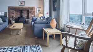 Ein gemütliches Wohnzimmer mit Kamin aus Backstein, einer Couch, einem Holztisch und Blick durch ein großes Fenster.