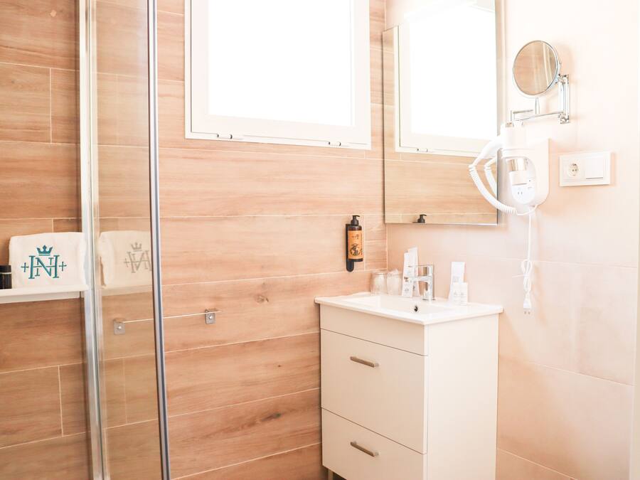 Un bany amb un espai de banyera de vidre, un lavabo blanc amb calaixos, un mirall i una prestatgeria amb productes de tocador.