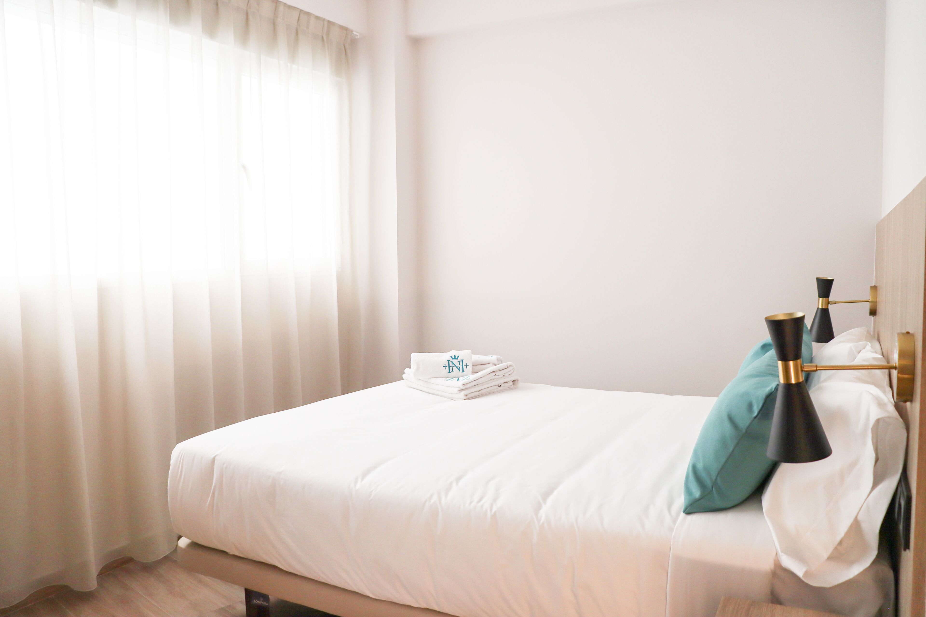 A neatly made bed with a folded towel and a teal pillow, a bedside lamp, and a window with sheer curtains.