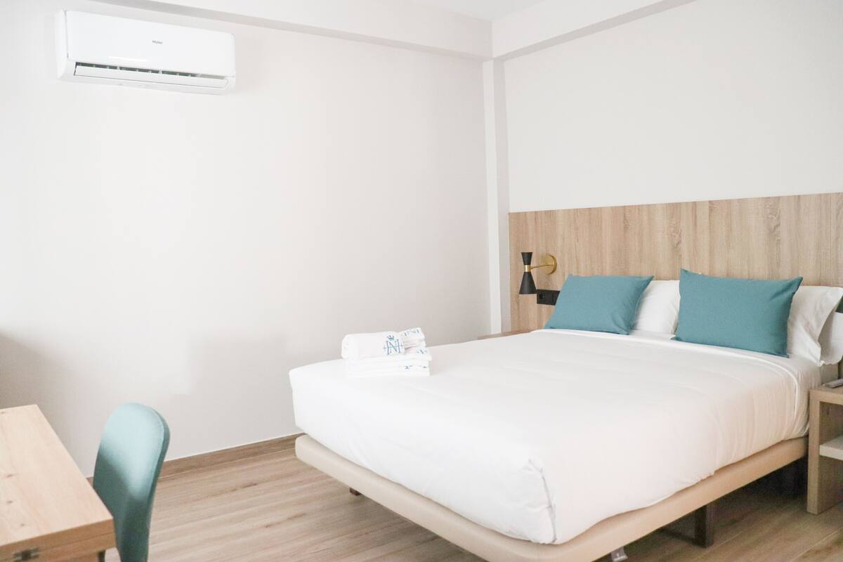 A hotel room with a bed, a wooden headboard, a nightstand, a desk, and an air conditioning unit.