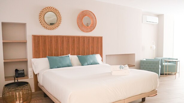 Un dormitorio ordenado con cabecera de madera, cama con ropa de cama blanca y turquesa, dos espejos en la pared, una mesita de noche con lámpara y una pequeña zona de estar con una silla y una mesa.