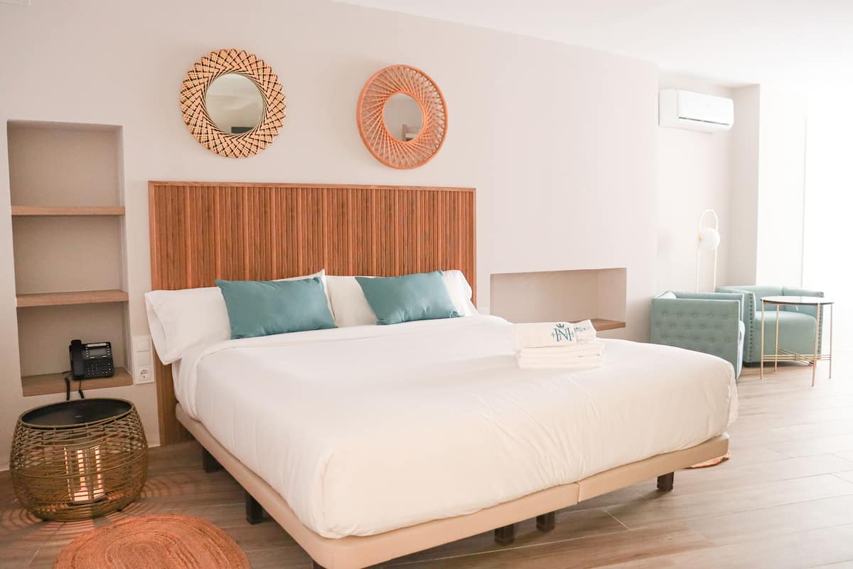 A neatly arranged bedroom with a wooden headboard, a bed with white and teal bedding, two wall mirrors, a bedside table with a lamp, and a small seating area with a chair and table.