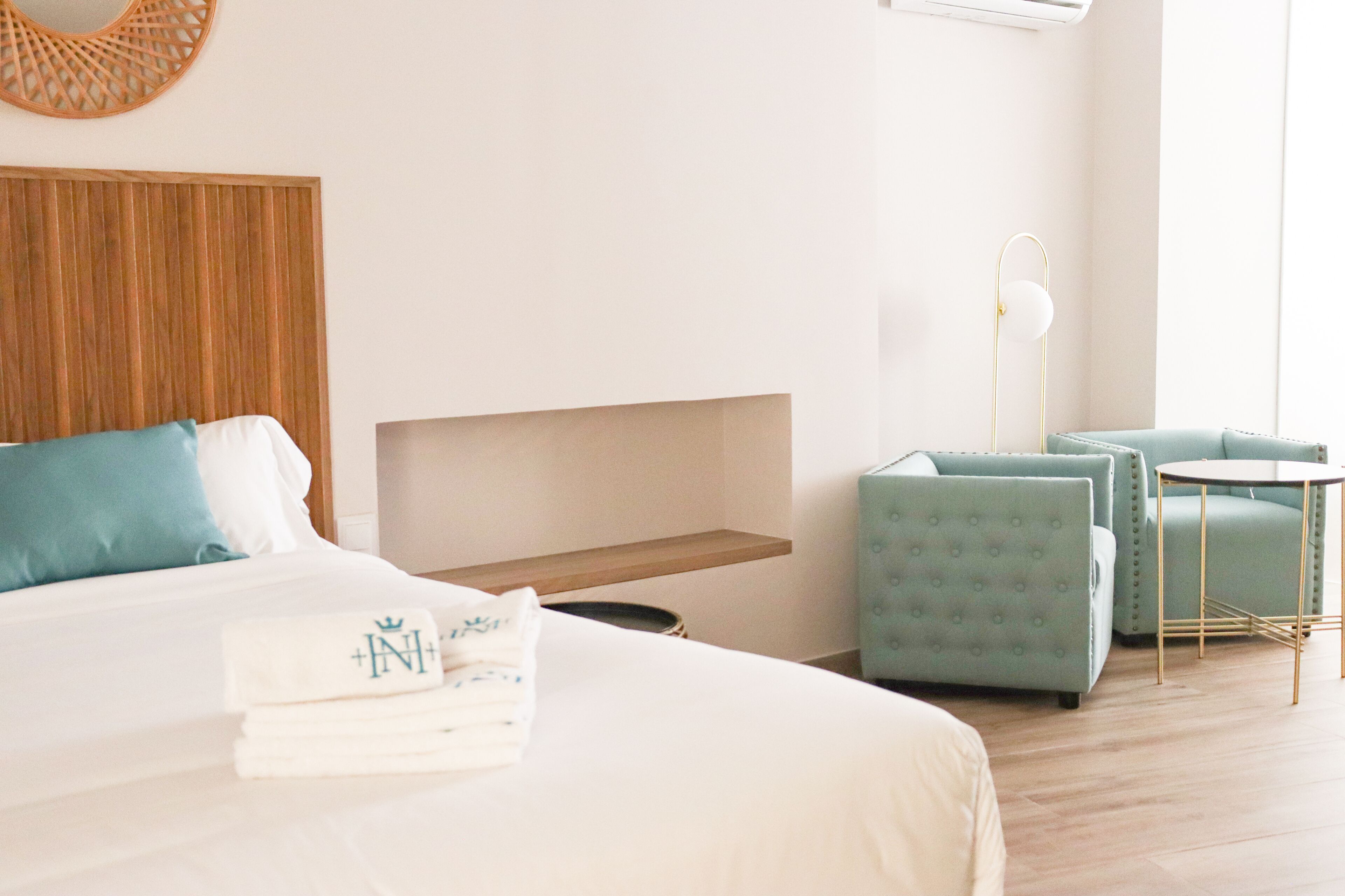 A neatly made bed with a pillow and a folded towel, a small bedside table, a wall-mounted shelf, a round table with a chair, and a floor lamp.