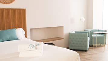 Una cama bien hecha con una almohada y una toalla doblada, una mesita de noche pequeña, un estante en la pared, una mesa redonda con una silla y una lámpara de pie.