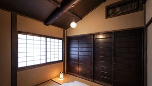 A traditional Japanese room with tatami flooring, a shoji screen window, a dark wooden headboard, and a hanging light fixture. - Rinn Shijo Machiya (Kyoto)