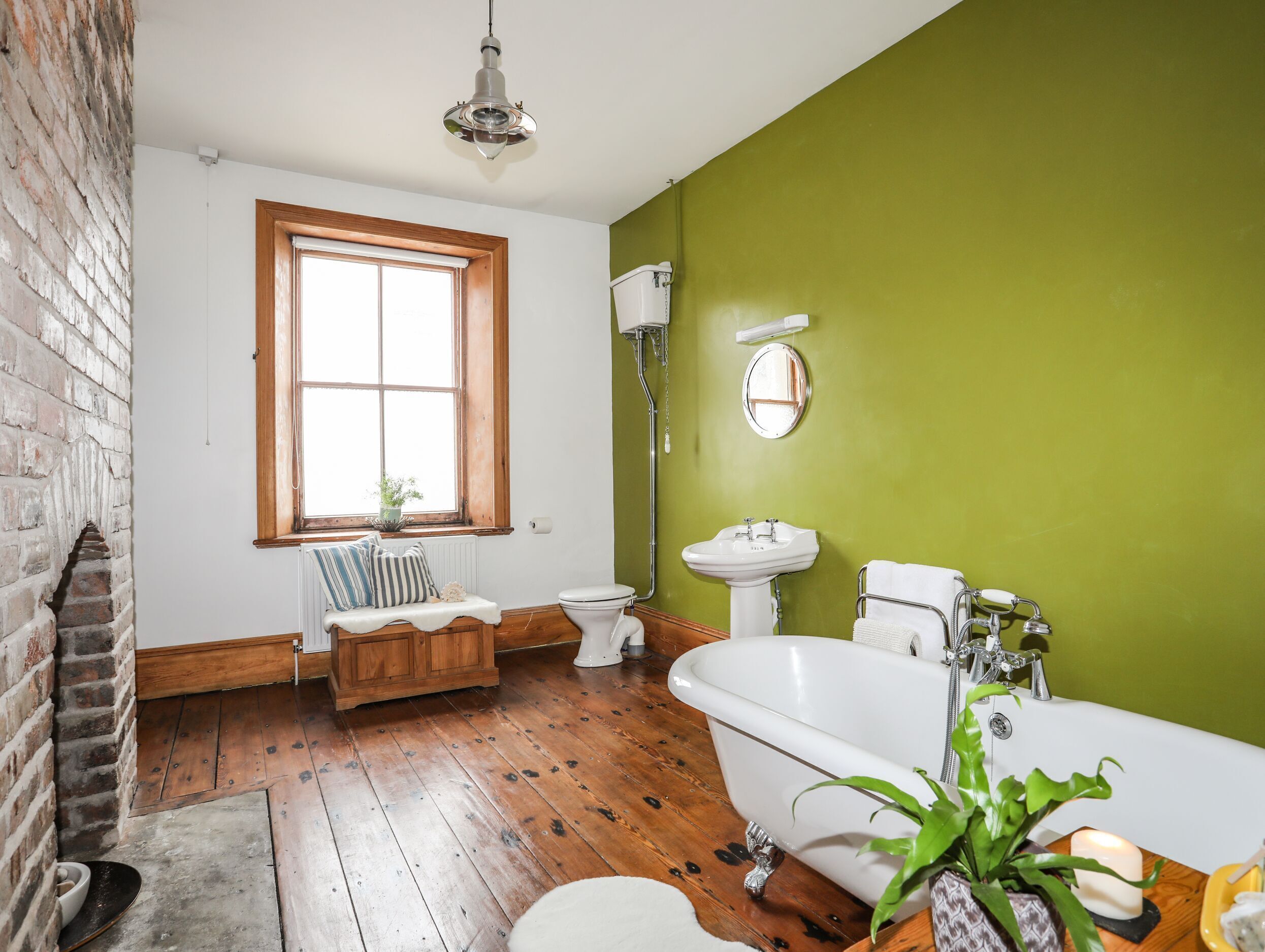 Un baño con piso de madera, una bañera blanca, un lavamanos blanco y un inodoro. Hay una ventana con marco de madera y una planta en el alféizar.