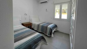 Une petite chambre propre, avec deux lits simples, une fenêtre avec des stores et un appareil de climatisation fixé au mur.