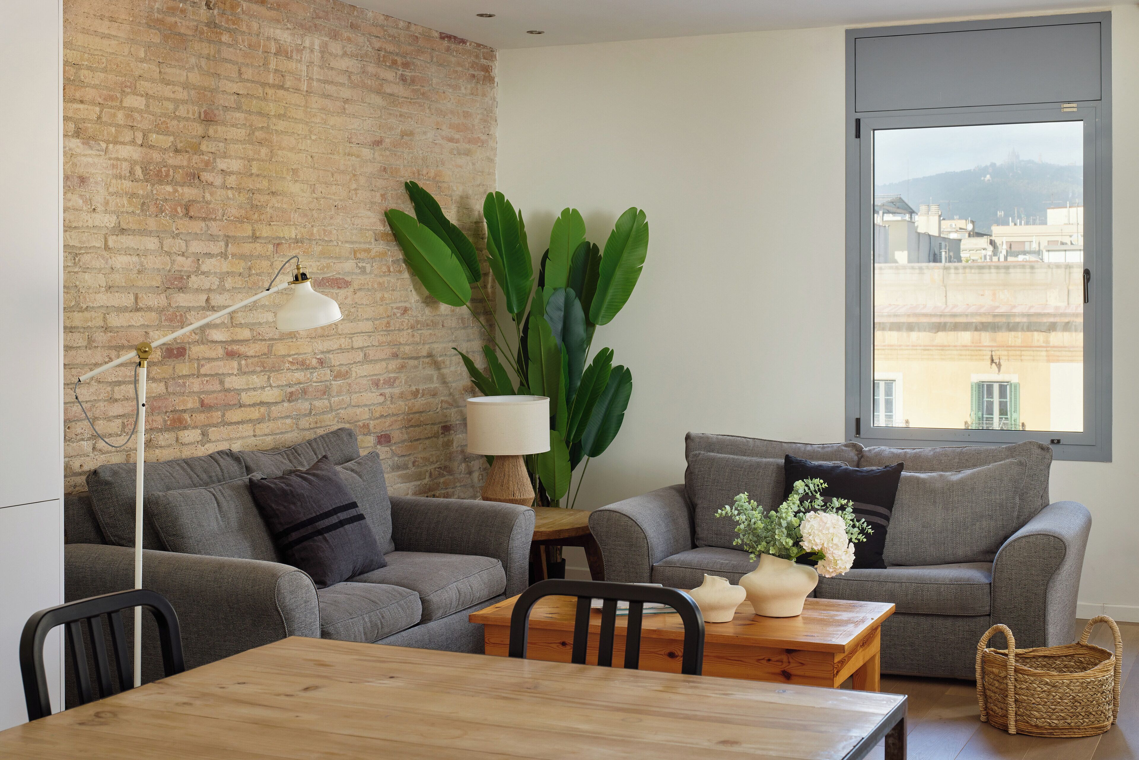 Een moderne woonkamer met een bakstenen muur, grijze banken, een houten tafel en een grote plant.
