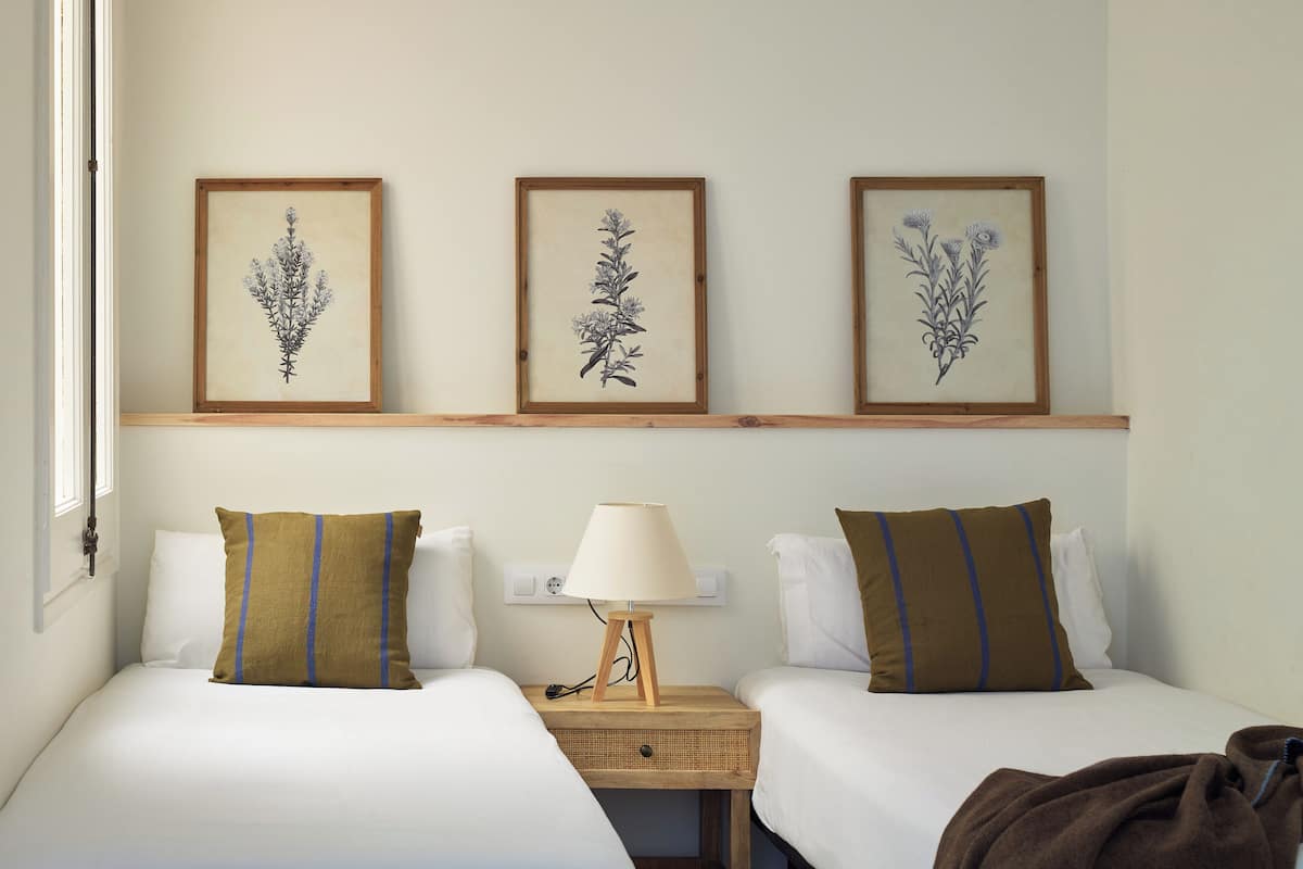 A hotel room with two beds, a bedside table with a lamp, and three framed botanical illustrations on the wall.
