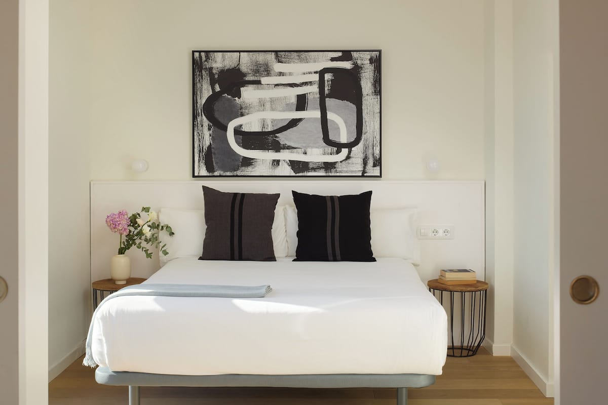 A neatly made bed with a white duvet, two black pillows, and a small bedside table with a book and a vase of flowers.