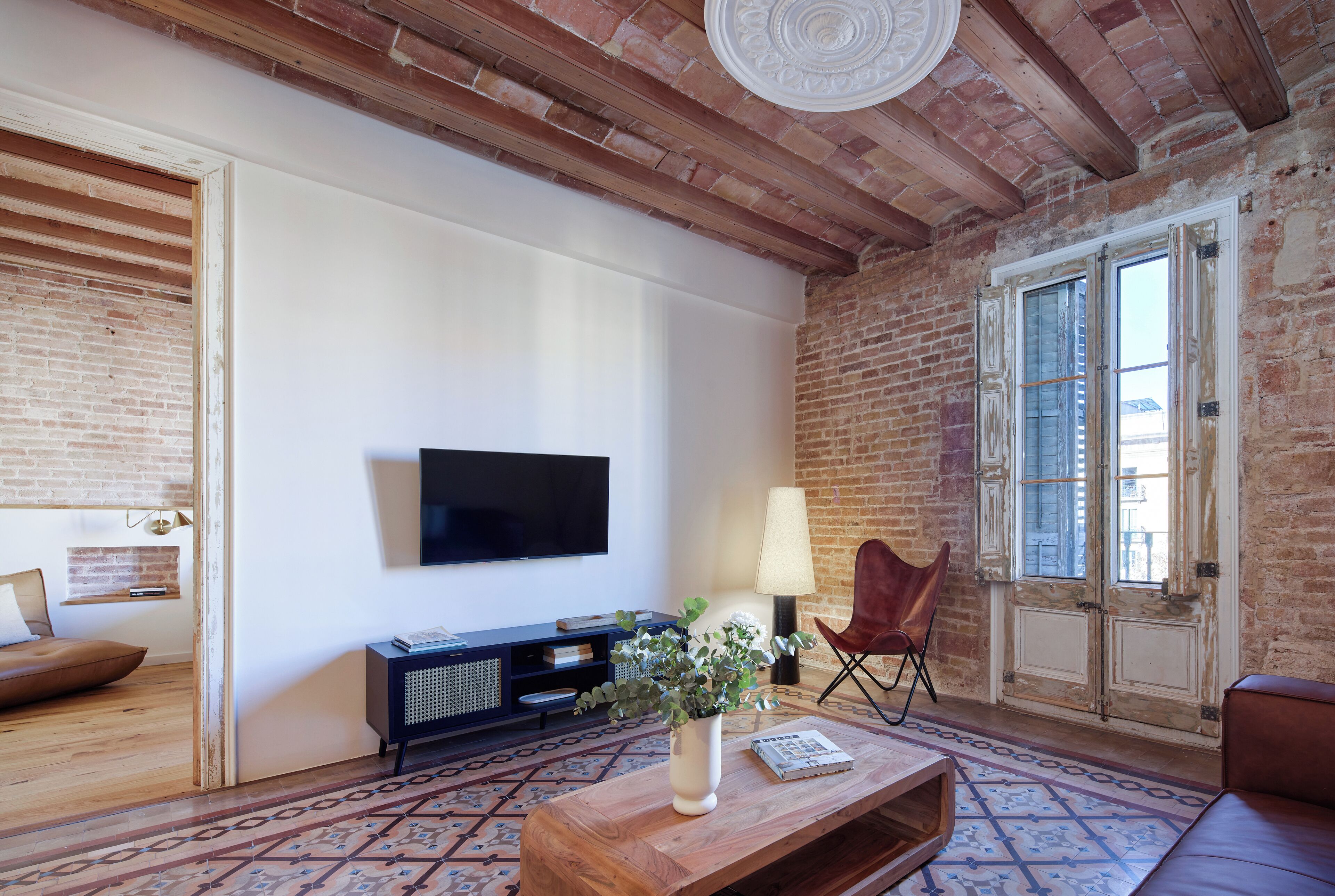 Un salon avec des murs en briques apparentes, un téléviseur à écran plat fixé au mur, une table basse en bois sur laquelle est posé un vase de fleurs, un tapis à motifs et un canapé en cuir.